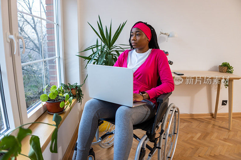 坐在轮椅上沉思的黑人妇女在使用笔记本电脑时透过窗户看