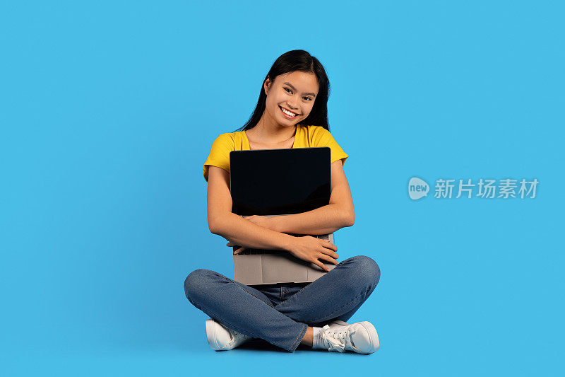 快乐的千禧亚洲女性在黄色t恤坐在地板上，抱着笔记本电脑，孤立在蓝色背景