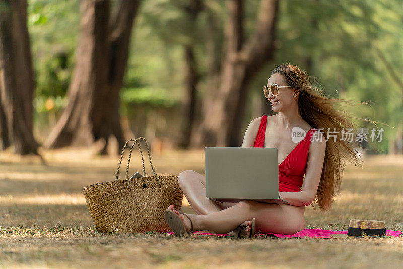 美丽的白人女性性感的女人在红色泳装比基尼戴着太阳镜在她的电脑笔记本电脑上工作，坐在海边的树林里放松