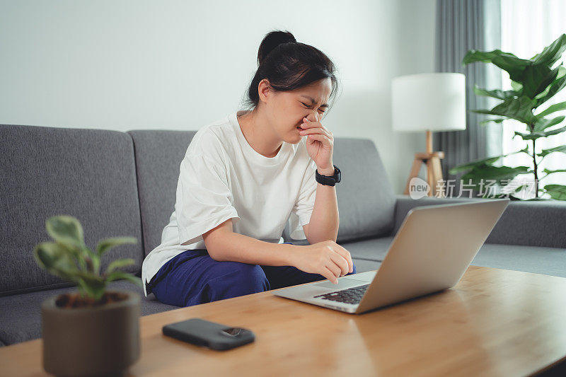 一名亚洲女性坐在家里客厅的沙发上，用笔记本电脑工作，喉咙痛，发烧。