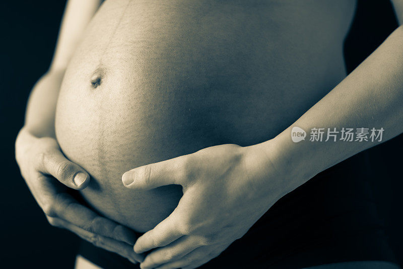 特写:穿着内衣的孕妇用手捧着赤裸的圆肚子。怀孕的最后一个月，第36周。侧角视图。黑色背景。局部塑身。