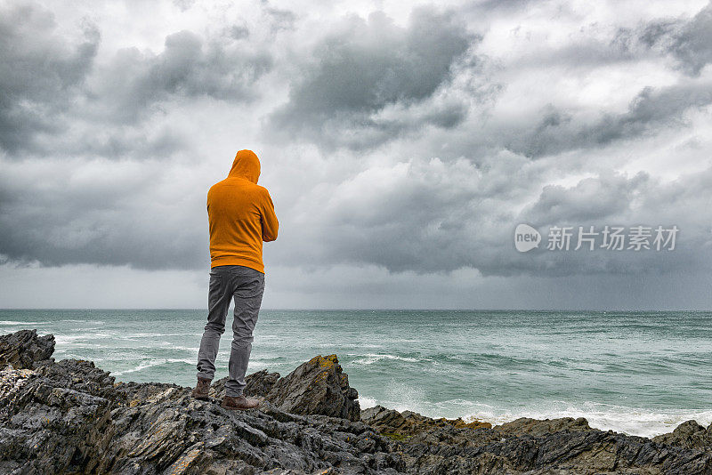 一个人站在海边的岩石上，俯视着阴沉沉的天空。