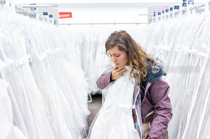 年轻女子在精品折扣店试穿蕾丝婚纱礼服，许多白色服装挂在衣架排