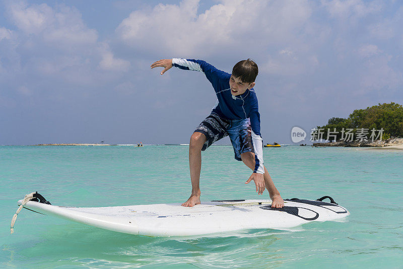 在马尔代夫古利岛的沙滩比基尼海滩上，一个小男孩在冲浪板上对着蓝天保持平衡
