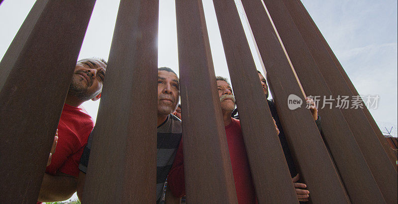 四个不同年龄的拉美裔男子(在墨西哥这边)透过墨西哥和美国之间的钢条边境墙凝视着美国(从美国这边拍摄)