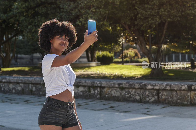 一个非洲裔美国女孩在阳光下用手机自拍，在户外公园散步