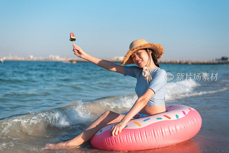 一个女人坐在沙滩上吃着西瓜，同时坐在一个漂浮的甜甜圈上