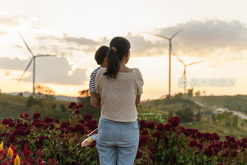 亚洲母亲抱着她可爱的儿子在风力涡轮机能源农场玩风车风力涡轮机探索户外学习可持续能源。儿童户外学习活动体验环保可持续的生活方式理念。