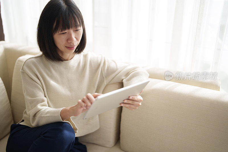 在特写镜头中，一位亚洲女性坐在沙发上，她的手正在使用电子平板电脑，同时在家工作，阳光温柔地照在她身上