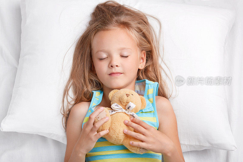 可爱的小女孩晚上做美梦，抱着泰迪熊，穿着睡衣，睡在卧室舒适的床上。有心爱玩具的小孩可以休息。童年的概念