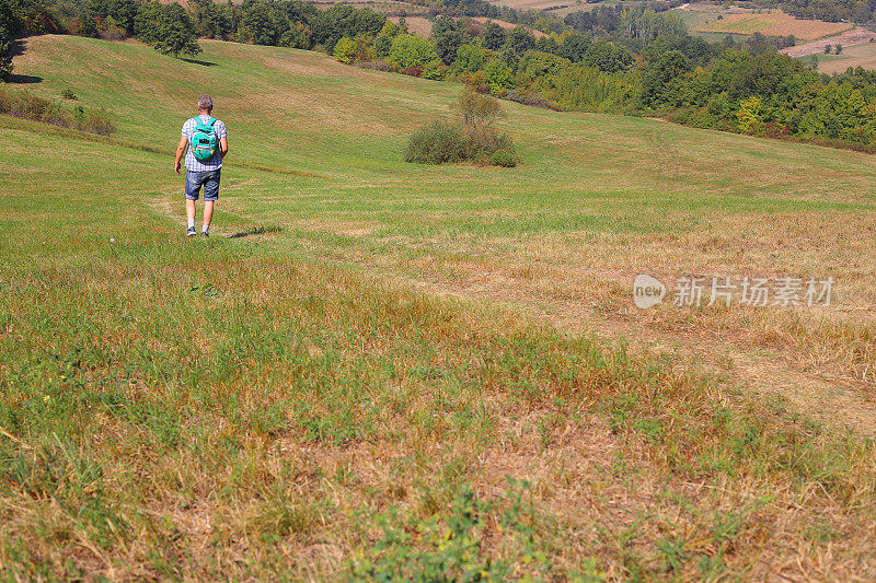 成年徒步旅行者徒步走过乡村的泥土路，穿过乡村自然的绿色草地