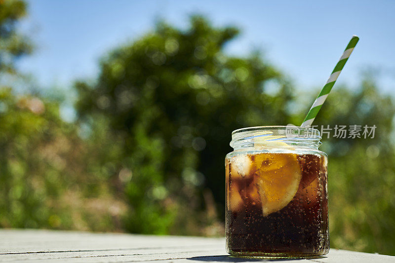 在一个炎热的夏天，花园的桌子上放着一个盛满可乐、冰块和一片柠檬的玻璃瓶。