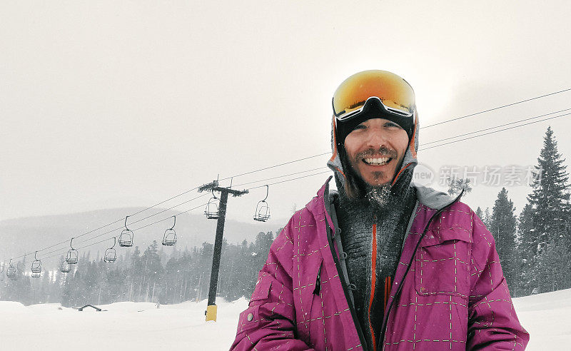 在科罗拉多州博尔德附近的埃尔多拉滑雪度假村，一个30多岁的滑雪者穿着全套的冬季装备和护目镜，在他身后的滑雪缆车上对着镜头微笑