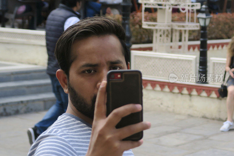 在拉贾斯坦邦的乌代普尔城宫殿外，年轻英俊的印度男子用手机相机自拍，拿着手机看手机屏幕自拍，印度男子头发梳到后面，棕色胡子，t恤，手镯