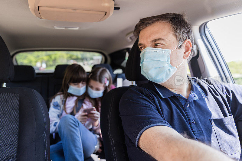 一个戴着外科口罩的男人开着车，看着他的女儿们在后座上玩