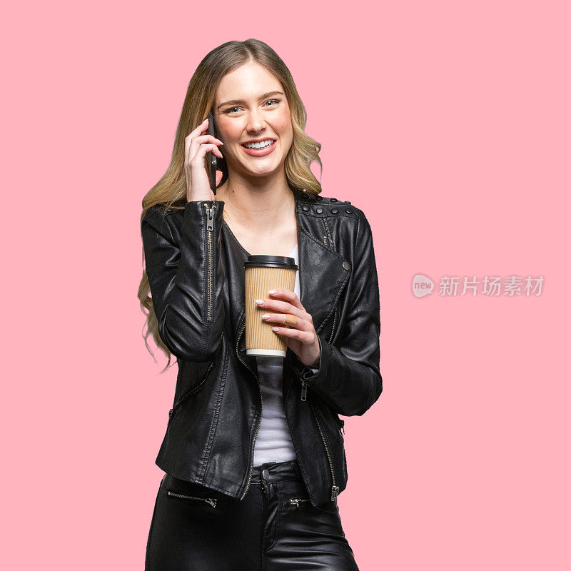 z一代年轻女性穿着裤子站在有色背景前，拿着咖啡杯，使用手机
