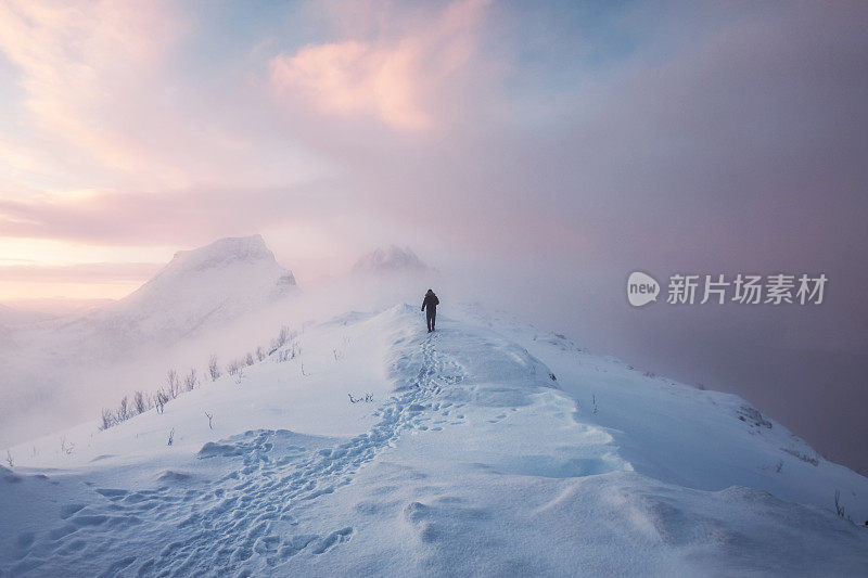 一名登山运动员，在暴雪的日出时分，踏着脚印在雪山和五彩的天空上行走