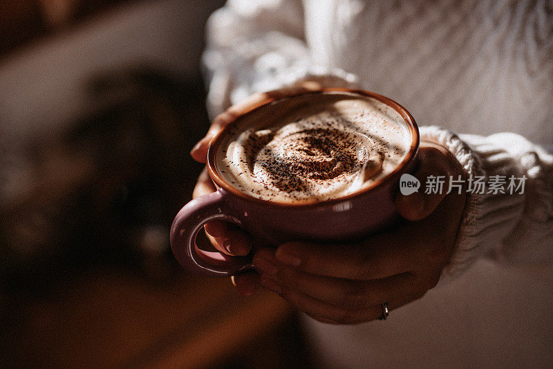 一个陌生的年轻女子，穿着白色的冬季套衫，手里拿着奶油咖啡，站在窗边