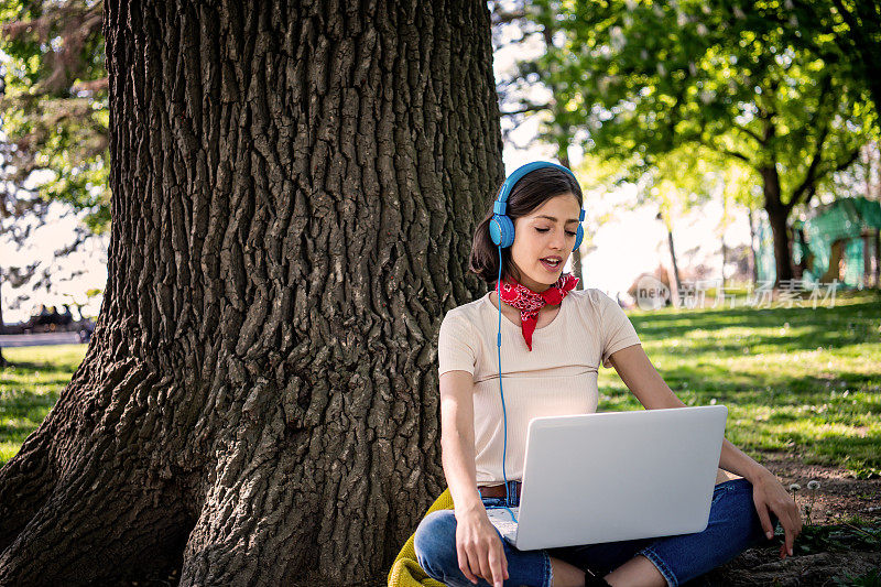 肖像的漂亮的年轻女子坐在绿色的草地上，双腿交叉在夏天的一天，而使用笔记本电脑和无线耳机进行视频通话