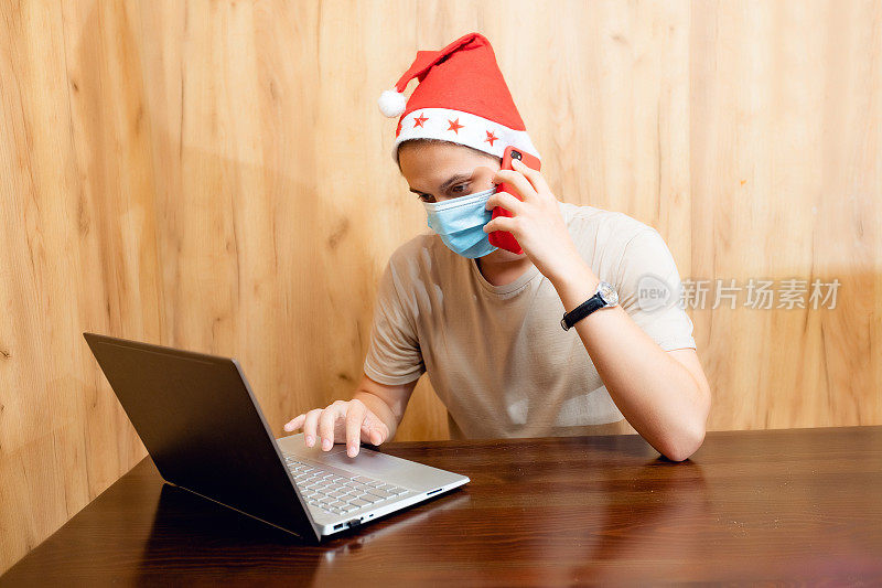 圣诞男性肖像，戴着红色圣诞帽和面部保护口罩的男子在大流行冠状病毒期间使用笔记本电脑进行视频通话，新年概念。有人在用手机打电话