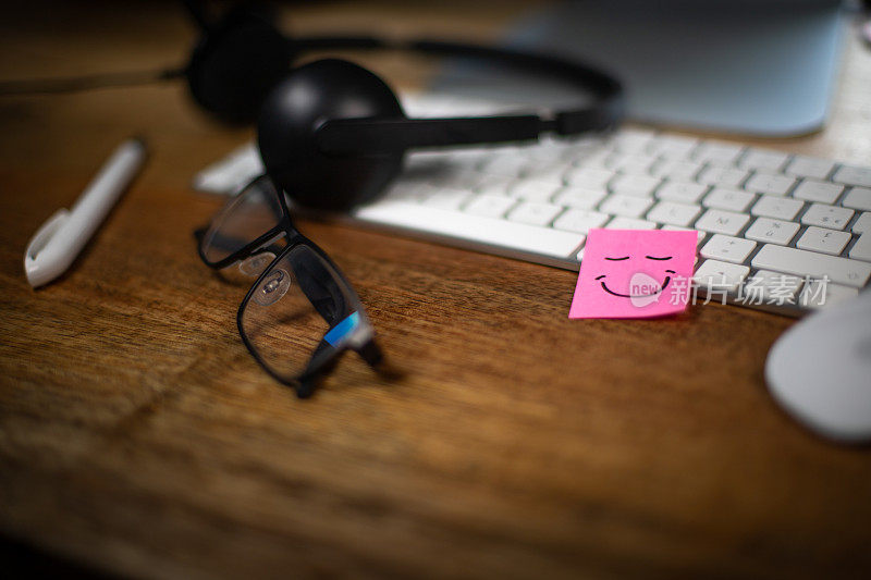 工作中幸福和幸福的象征。配备电脑、眼镜和耳机麦克风的工作站。键盘上贴着一张带微笑的粉色便签。