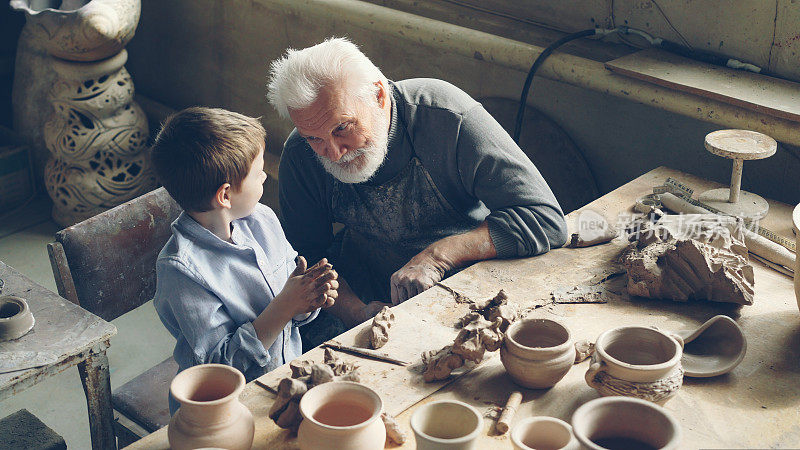 好奇的小男孩在作坊的桌子旁和他的专业陶工爷爷一起揉泥聊天。家庭传统，陶器和童年观念。