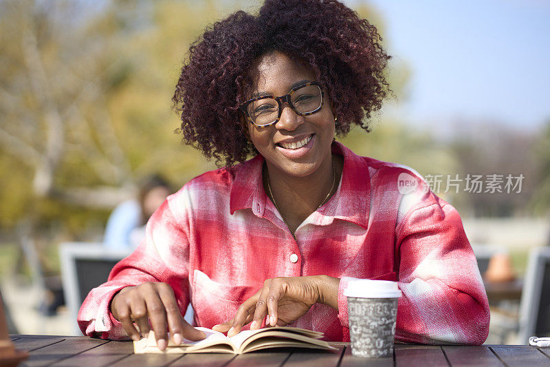 年轻的黑发拉丁非洲头发，手里拿着一本书，拿着一杯要外卖的咖啡，在一个阳光明媚的早晨，坐在外面摆出一副若有所思的姿势。