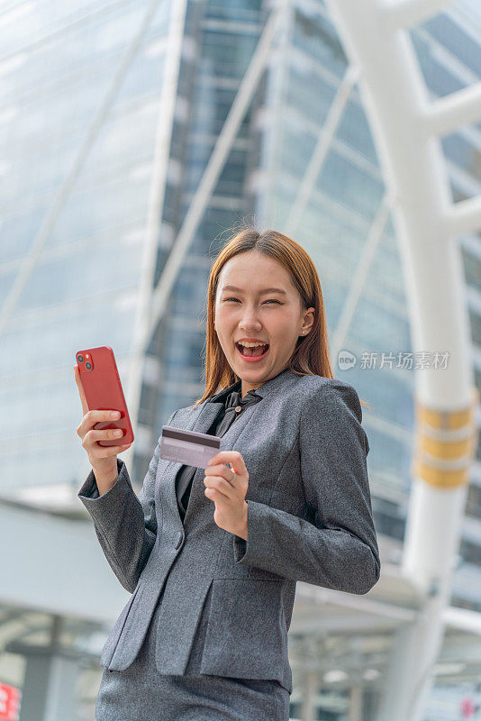 一名年轻的亚洲女商人穿着灰色的办公套装，在商业区用她的智能手机使用信用卡进行在线交易，她微笑着自信