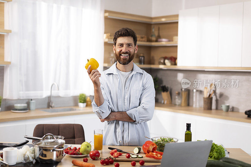 微笑的成年欧洲大胡子男性拿着胡椒，在现代厨房内部用有机蔬菜准备沙拉