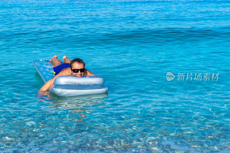 黝黑的黑发男子在气垫上放松，在平静清澈的蓝色大海上摇摆。