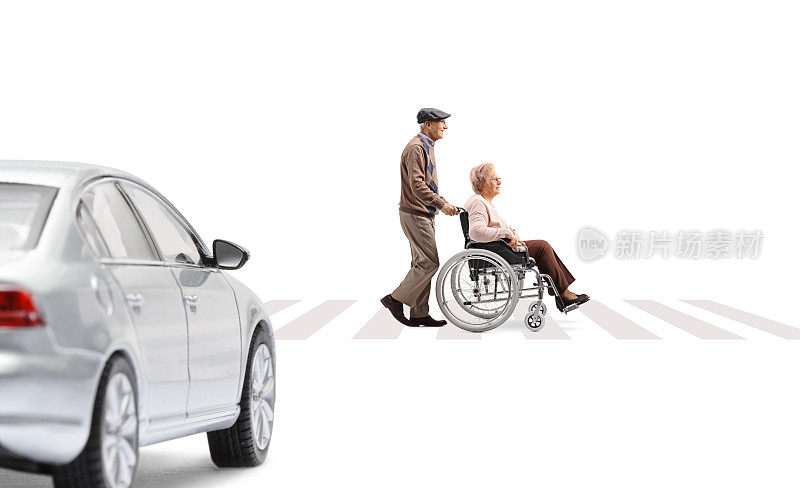 一名老年男子推着一名坐在轮椅上的老年妇女在人行横道和等待的汽车的全长侧影