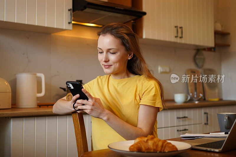 一个穿着黄色t恤、头发蓬松的女人拿着羊角面包和笔记本电脑坐在桌边，正在用手机打字。远程学习和工作理念
