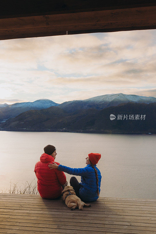 在挪威的日落时分，幸福的夫妇和狗在避难所欣赏冬季峡湾的美景