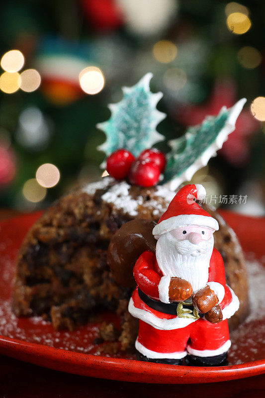 圣诞老人雕像站在自制的红盘子上的特写图像，用冬青叶和红浆果装饰的圣诞布丁，缺失的部分，节日的圣诞树和照亮的仙女灯背景，重点在前景