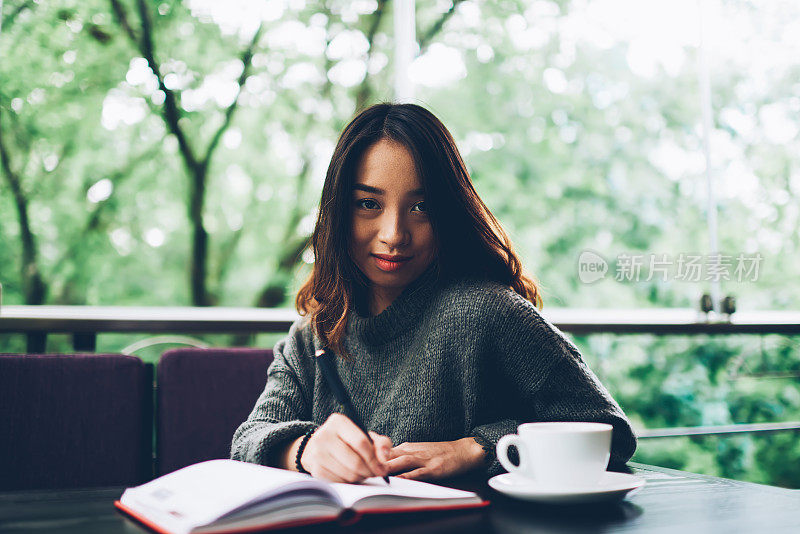 肖像迷人的年轻亚洲女人在舒适的咖啡馆阳台上做笔记在日记，集中的女性经理计划一天，在笔记本上制定时间表在咖啡休息时间