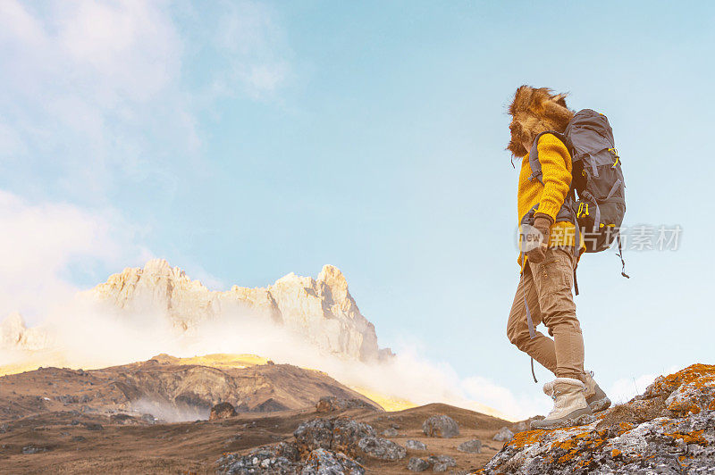一个背着背包的女孩戴着墨镜，戴着一顶大大的北方皮草帽，背着一个背包，站在一块岩石上，看着隐藏在岩石中的悬崖