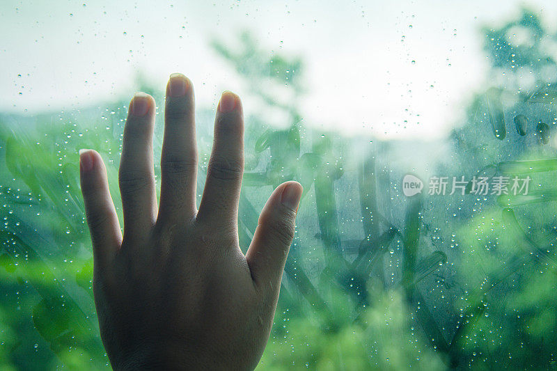 特写镜头多雨的心情。下雨天有人碰窗户。在阳光和树的背景下，雨滴落在窗户玻璃表面。下雨天的孤独。软焦点
