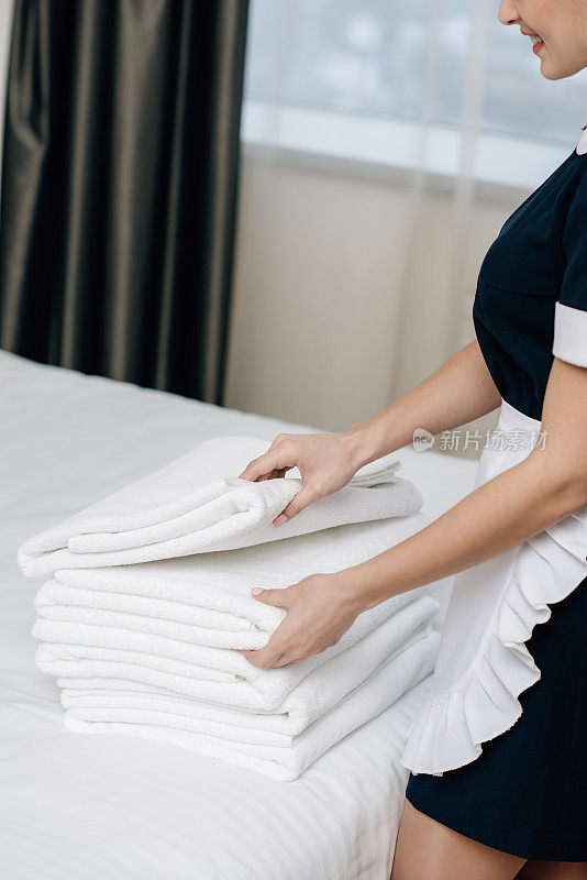 酒店套房的床上放着一堆干净毛巾，穿着制服的女保洁员微笑的镜头