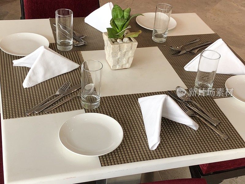 白色方形餐厅餐桌的形象，为早餐，午餐或晚餐照片与四个棕色的餐垫，银餐具刀，叉和勺子，盘子，水杯，米色和白色主题折叠布餐巾，红色椅子