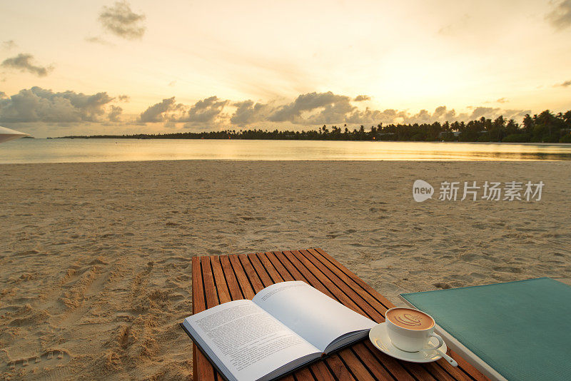 在日落海滩背景的木桌上，打开一本书和一杯咖啡