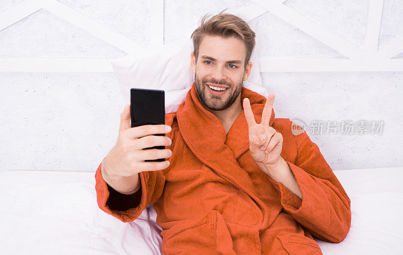 有问题的明星。快乐的男人在床上用智能手机自拍。帅哥笑着用V手势自拍手机。在卧室享受自拍