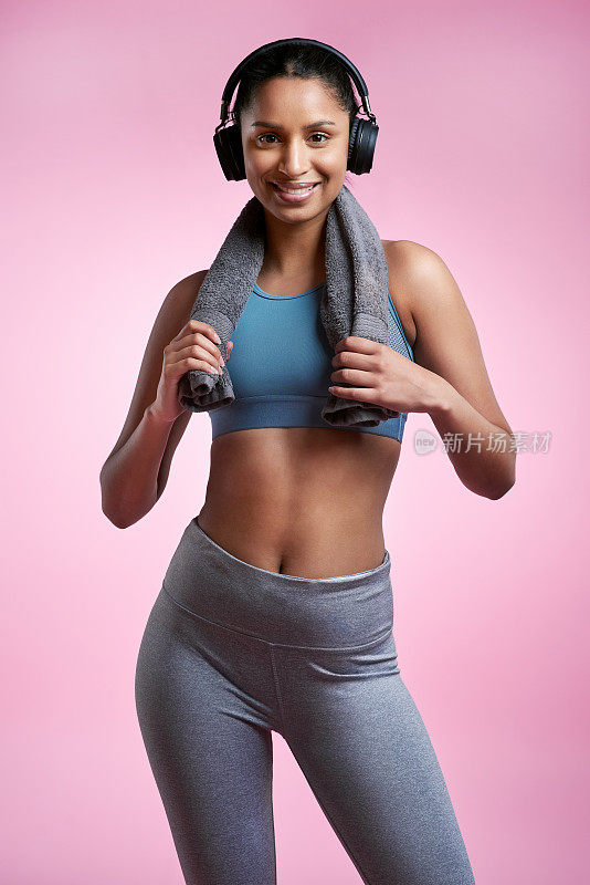 裁剪肖像的一个迷人和运动的年轻女子戴着耳机和摆姿势与一条毛巾在工作室的粉红色背景