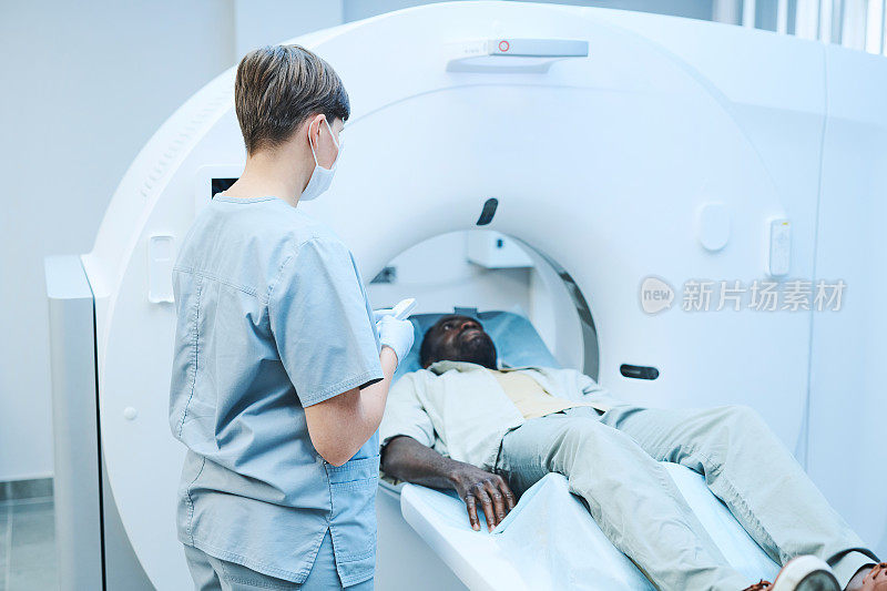 年轻护士戴着口罩在MRI扫描仪控制面板上按下启动按钮，同时在临床中执行操作