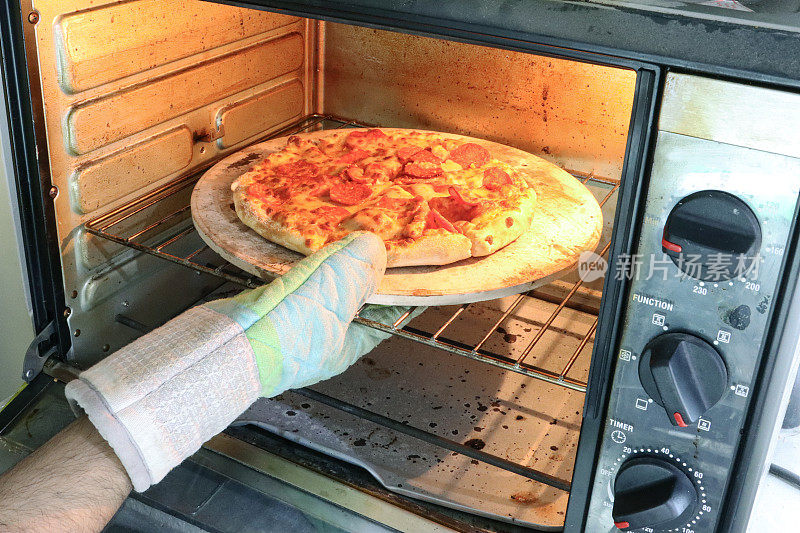 照片中，一个面目全非的人戴着烤箱手套，从热烤箱中取出自制的意大利辣香肠披萨，在披萨石头上烤得脆脆的披萨，磨碎的马苏里拉奶酪和意大利辣香肠薄片，焦点放在前景上