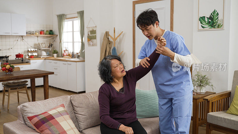 富有爱心的亚洲男性个人理疗护理助理，帮助中风老年女性患者在家中客厅进行康复训练。他慢慢抬起她疼痛的手臂