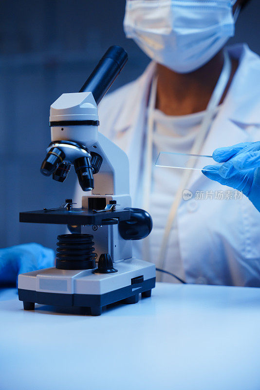 一个非裔美国医生的手，戴着防护手套，手里拿着显微镜载玻片，在实验室里的一张装有显微镜的桌子上