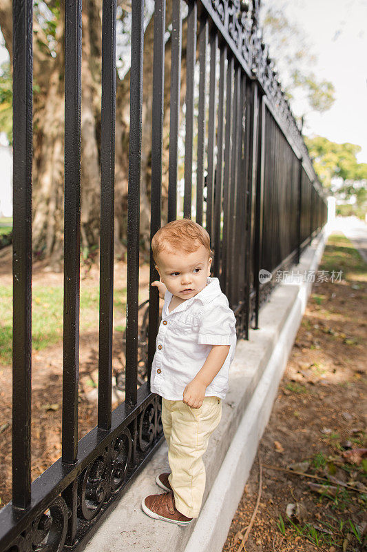 一个可爱的一岁古巴裔美国男婴，穿着白色亚麻短袖衬衫，淡黄色裤子和棕色鞋子，站在佛罗里达州棕榈滩的一条湖上，紧紧抓住黑色栏杆