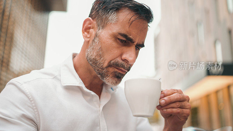 阴云密布，成熟的商人，留着整齐的胡子，穿着白衬衫，坐在咖啡桌旁，喝着咖啡，用智能手机打字