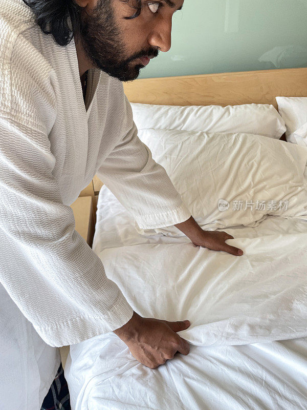 特写图像的印度男子穿着白色毛巾浴袍制作酒店双人床与木材和玻璃床头板，白色被褥被推翻，堆枕头，重点在前景
