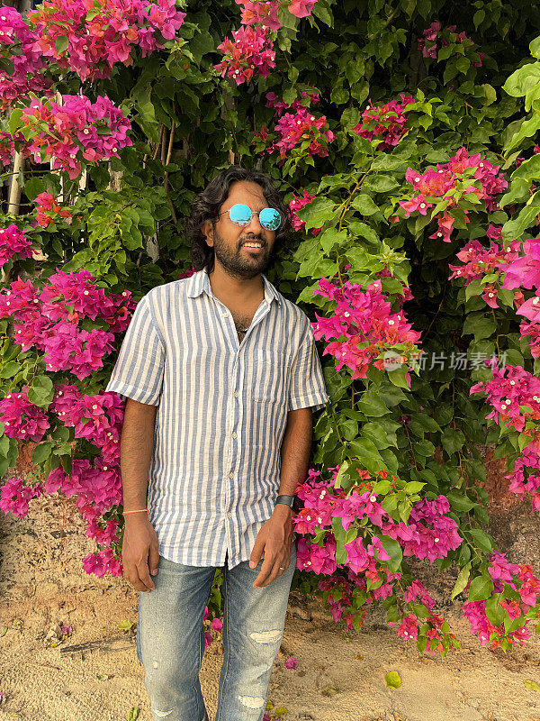 照片中，一名印度男子戴着彩色的镜面太阳镜，站在粉红色的九重葛花的背景前，这些花生长在葡萄藤上，就像不同寻常的热带花园树篱一样，这是前景的焦点